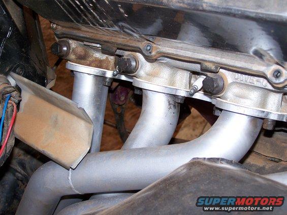 Close-up of exhaust manifold pipes connected to engine block with bolts. 'DOVE-C' is visible