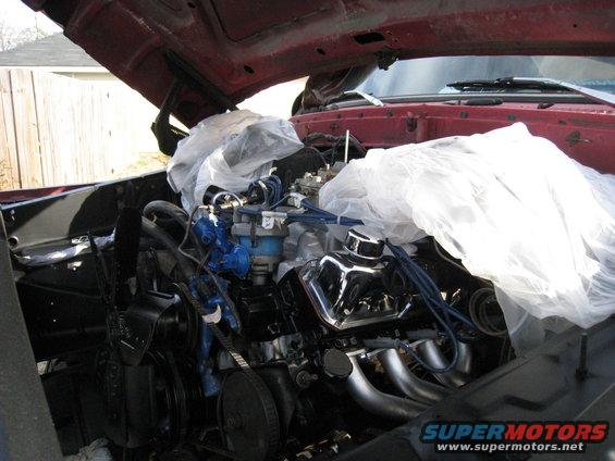 Engine bay with parts covered in plastic, showing chrome valve covers and exhaust headers