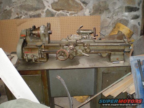 Metal lathe on a workbench in a stone-walled garage. The lathe is green and gray with various wheels and levers