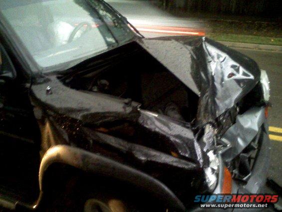Front end of a black SUV, heavily damaged after a collision. Hood is crumpled, and front bumper is torn