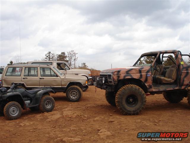 Four-door SUV, pickup truck with camouflage paint, and ATV parked on dirt