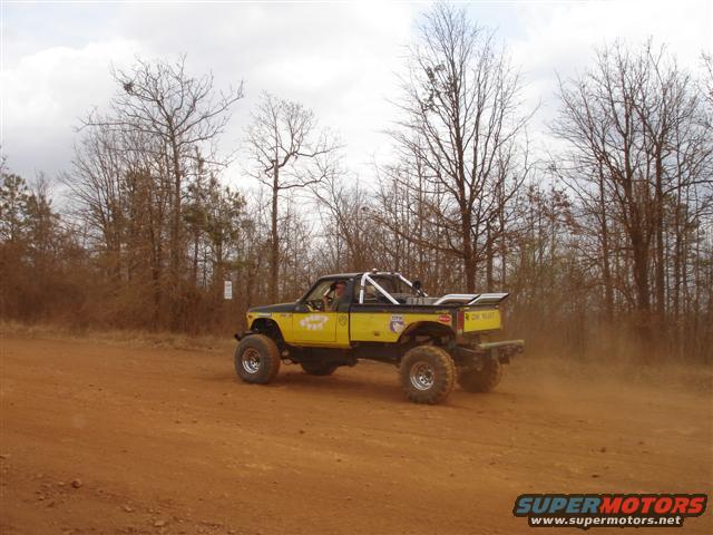Yellow pickup truck with large tires driving on a dirt road, kicking up dust. Has a roll cage and spoiler