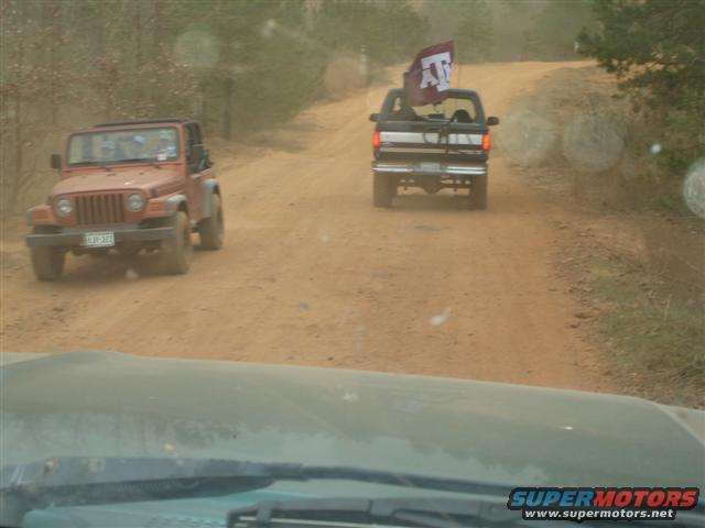 Orange Jeep and black pickup truck driving on a dirt road, pickup has a flag