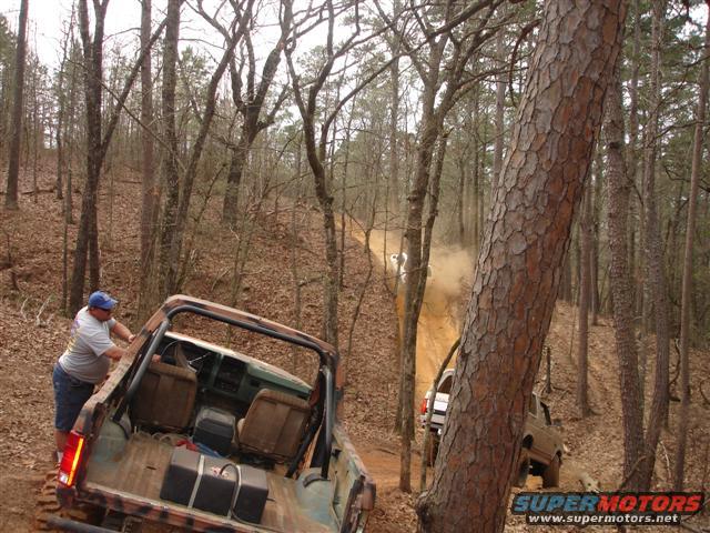 Off-road vehicles navigating a dusty dirt trail through a wooded area; one vehicle is in the foreground, being pushed by a person