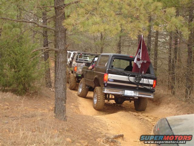 Black Ford Bronco with a maroon flag on the back, driving on a dirt road with other vehicles