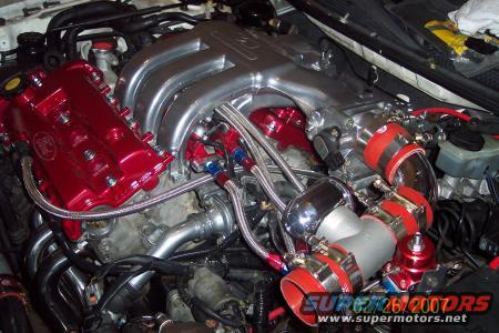 Engine bay with polished intake manifold, red valve covers, and custom exhaust headers. Supermotors.net logo visible