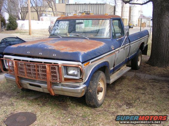 Rusty blue and silver Ford pickup truck with a large orange grille guard parked next to a tree.