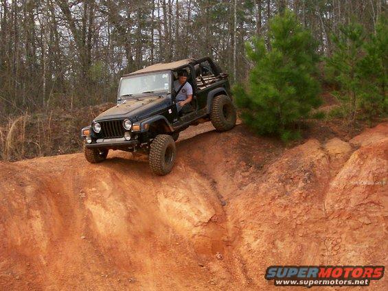 Black Jeep Wrangler TJ on a dirt hill, driver visible, soft top up, off-road tires