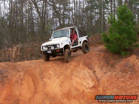 White Suzuki Samurai climbing a dirt hill with a driver wearing a red shirt