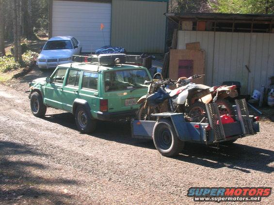 Green Jeep Cherokee with roof rack towing a trailer with two dirt bikes
