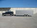 1995 Ford C-350 (Centurion Conversions) - A dark green SUV tows a large silver enclosed trailer in front of a concrete building.