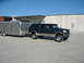 1995 Ford C-350 (Centurion Conversions) - A dark green Ford Bronco, likely from the mid-1990s, is parked next to a silver enclosed trailer under a clear blue sky.