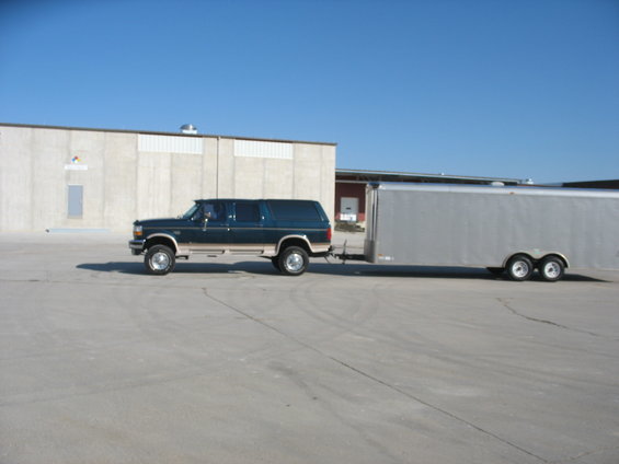 Dark green Ford SUV with tan trim towing a silver enclosed trailer. It's parked on pavement in front of a building