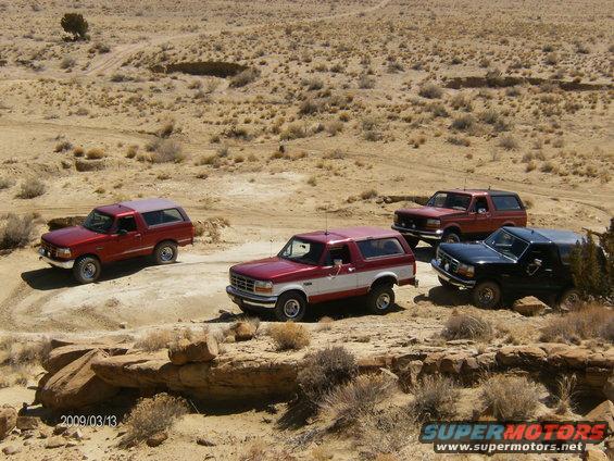 Four Ford Bronco SUVs parked in a desert landscape. The red and white Bronco in the foreground is a 1993 model