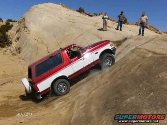 Red and white Ford Bronco climbing a rocky incline with people watching