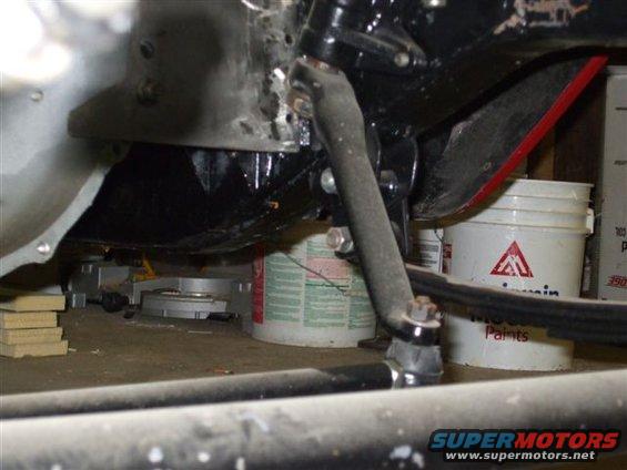 Close-up of a steering linkage component, possibly a pitman arm, with buckets and shop items in the background