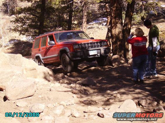 Red Jeep Cherokee XJ navigating rocky terrain with two people watching