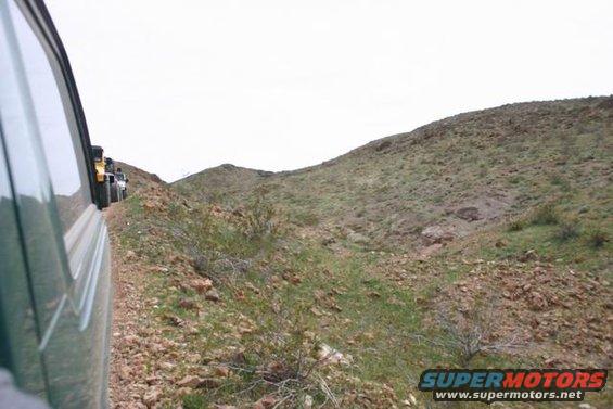 Line of off-road vehicles driving on a narrow dirt path through a rocky, arid landscape