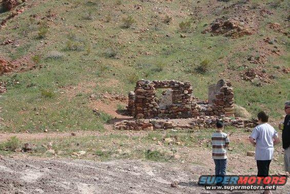 Stone ruins stand in a dry, grassy landscape with three people observing