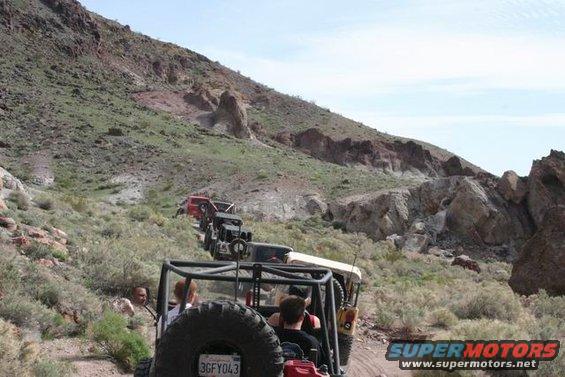 Line of off-road vehicles driving up a rocky mountain trail under a clear sky. License plate visible on the lead vehicle