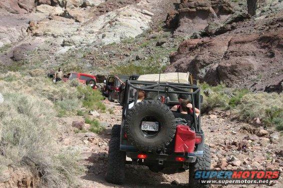 Several off-road vehicles driving up a rocky canyon trail. The lead vehicle has a spare tire mounted on the back