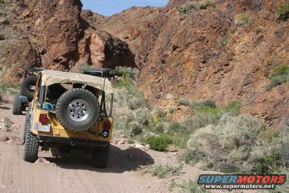 Yellow Jeep Wrangler with tan soft top, spare tire on rear, driving on dirt road