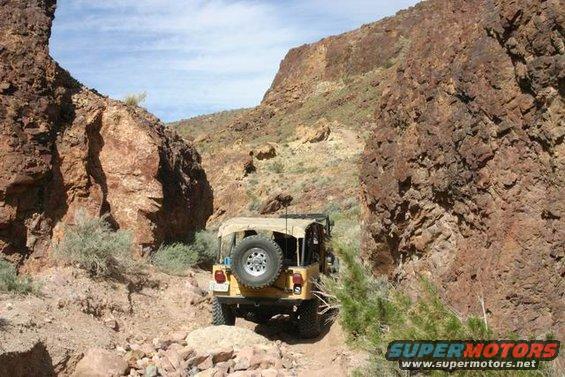 Yellow Jeep CJ-7 with a tan soft top driving on a rocky, narrow trail between large rock formations