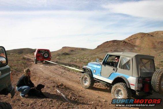 Blue Jeep being towed by a red Jeep on a desert trail, with a woman and dog watching