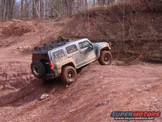 Mud-covered silver Hummer H3 SUV with a roof rack and spare tire on a dirt trail