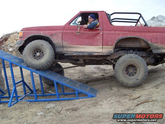 Red SUV with off-road tires on a blue ramp, driver visible, roll cage in rear
