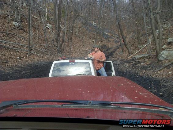 View from inside a red vehicle looking at a white SUV on a muddy off-road trail with trees and hills in the background