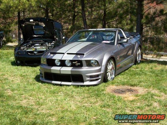 Gray convertible Mustang with racing stripes parked on grass, black Mustang with hood open behind it