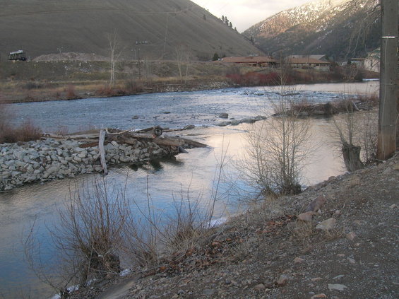 River flowing past a rocky bank with bare trees and distant buildings on a cloudy day.