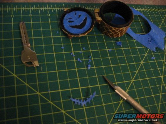 Craft project with a key, a circular object with a smiley face made of blue tape, and a hobby knife on a cutting mat
