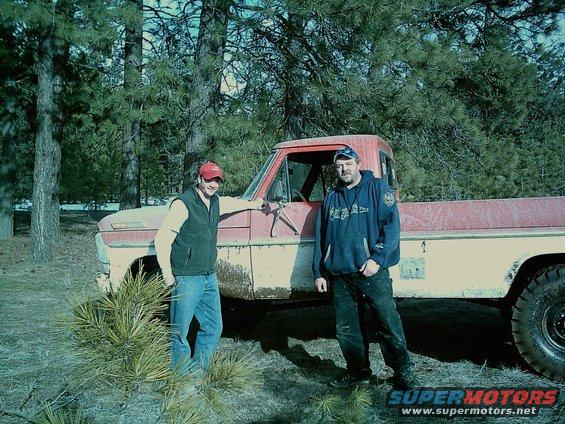 Two guys stand next to a red and white pickup truck in a wooded area