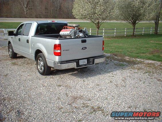 Silver Ford F-150 pickup truck parked on gravel, carrying a V8 engine in the bed. Trees and a white fence in background