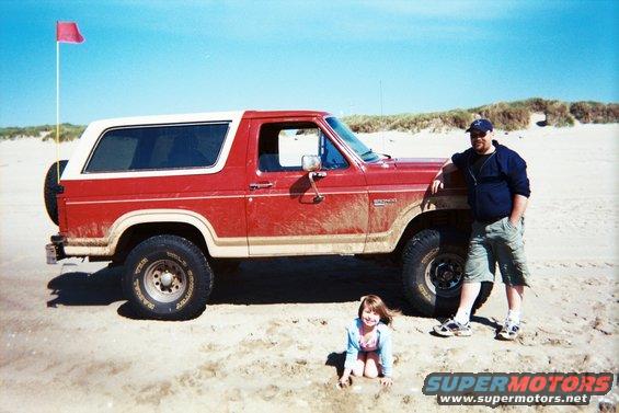 Man and girl by a red 1985 Ford Bronco on a sandy beach with dunes and a red flag