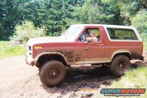 Red Ford Bronco with white top driving through mud, driver visible