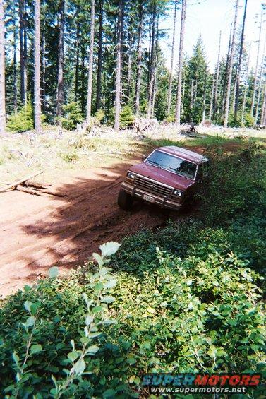 Red 1985 Ford Bronco driving off-road on a dirt path through a forest