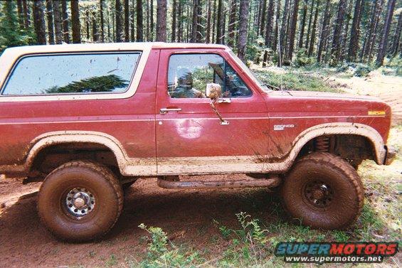 Side view of a red 1985 Ford Bronco covered in mud, driving on a dirt path in a forest
