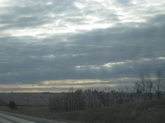 Overcast sky with sunbeams breaking through clouds over a rural landscape with dry grass and trees