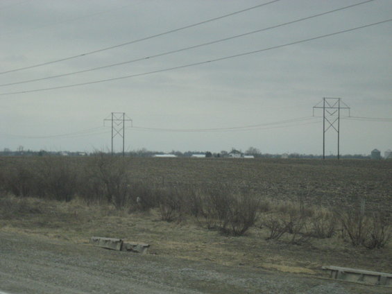 Power lines stretch across a cloudy sky over a barren field with distant farm buildings