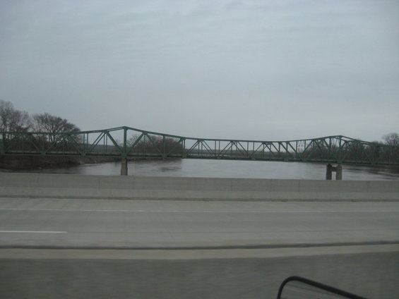 Green truss bridge over a wide, murky river under an overcast sky, seen from a highway