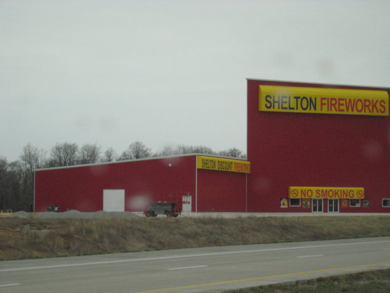 Two red industrial buildings, one with a large 'SHELTON FIREWORKS' sign, the other with 'SHELTON DISCOUNT FIREWORKS'. A 'NO SMOKING' sign is visible near the entrance of the larger building