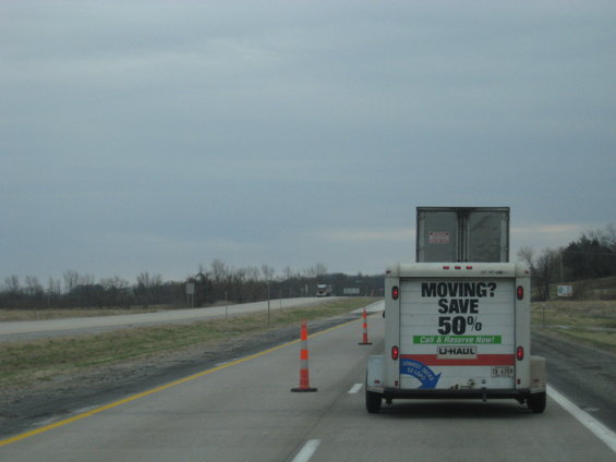 U-Haul trailer with 'MOVING? SAVE 50%' advertisement, followed by a semi-truck, on a highway