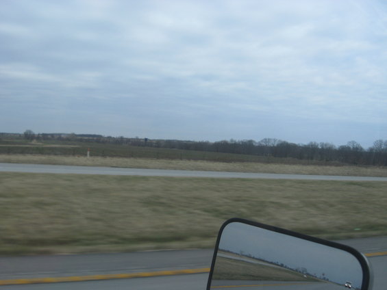 view of a highway with fields and trees under a cloudy sky, seen from a vehicle's side mirror