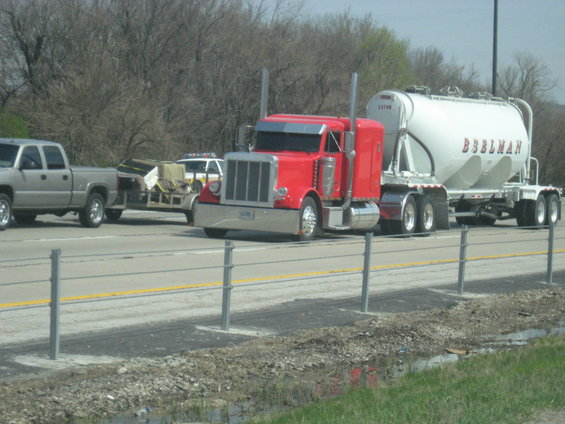 Red Peterbilt semi-truck with a white cement trailer, a silver pickup truck towing a trailer, and a white vehicle are on a highway