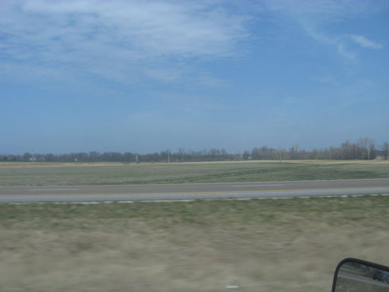 view of a wide, flat field under a blue sky with scattered clouds, seen from a moving vehicle