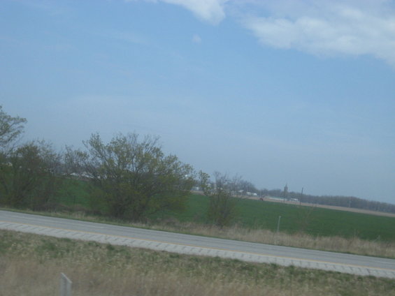 View from a car on a highway looking out at a green field and some trees under a blue sky with clouds