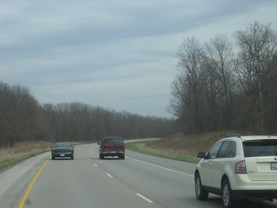 Several vehicles driving on a highway with bare trees on the side